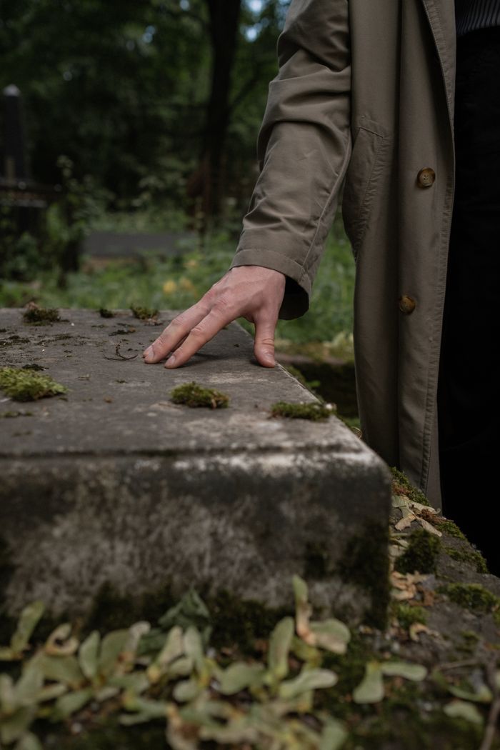 A hand gently touches a moss-covered tombstone in a tranquil cemetery setting.