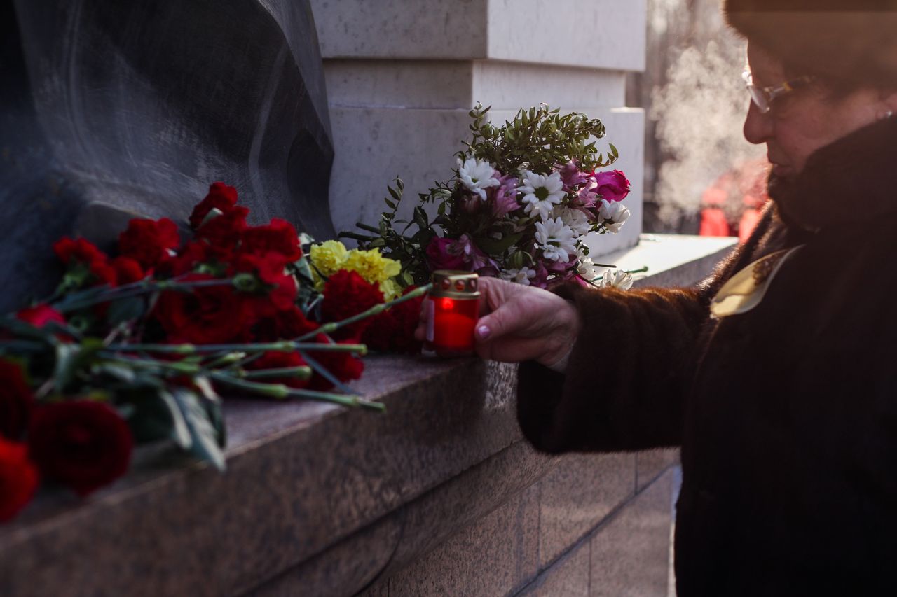 An elderly woman places flowers and a candle at a cemetery memorial site, reflecting homage and remembrance.